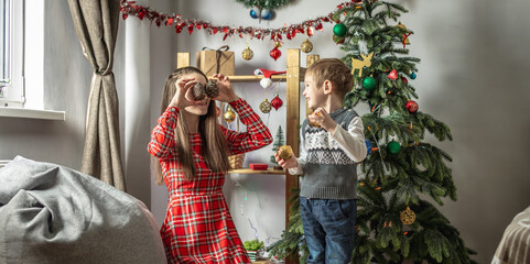 Young woman and her little son are decorating a Christmas tree together and fooling around and having fun. Concept of a festive atmosphere, preparation for the New year, family pastime