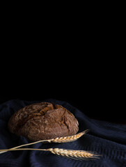 Round loaf of rye bread with a spike of wheat on a blue towel in a dark key on a black background, vertical photo, free space for text