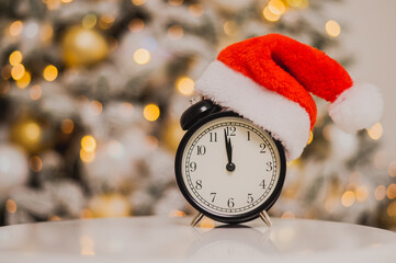 Alarm clock in a santa hat against the background of a christmas tree.