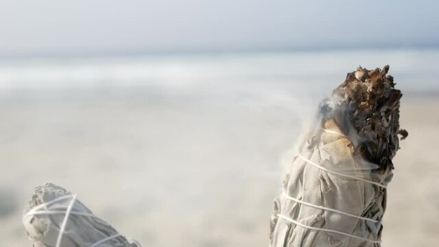 Dried white sage smoke, dry smudge stick burning, soft focus bokeh. Aroma smudging close up on ocean beach, aromatherapy meditation by sea water waves on sandy coast. Yoga, peace, harmony and ayurveda