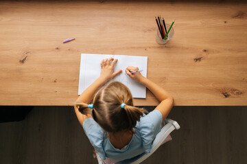High-angle view of unrecognizable primary little child girl doing homework sitting at home table by window. Smart preschool kid studying alone drawing with pen in bedroom. Homeschooling concept.