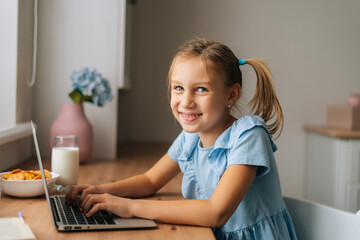 Side view of cheerful elementary child girl using laptop sitting at home table with snack by window, looking at camera. Portrait of little primary kid chatting having distance studying online.