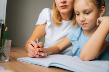 Close-up cropped shot of primary daughter doing homework with young mother sitting at home table by window. Pretty female tutor teaching elementary girl helping with lesson, selective focus.