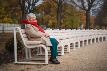 Senior woman sitting on bench in town park in autumn, looking at camera.
