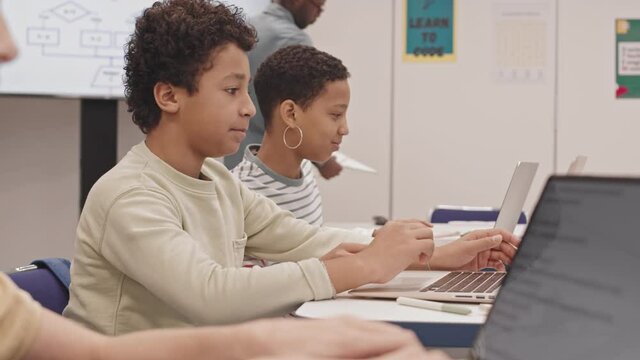 Side view of Black girl and Biracial boy smiling and talking, sitting at desks in classroom at IT lesson, using portable computers