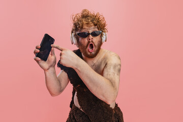 Portrait of man in character of neanderthal in headphones pointing at phone with shocked expression...