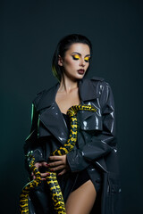 Naklejka premium Beauty woman python yellow snake around her neck on latex shiny raincoat. Yellow snake on the shoulders of girl. Beauty yellow eye shadow makeup, dark burgundy lipstick