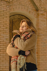 Blonde young woman in beige jacket and scarf wrapped herself against the background of brick building. Winter season. Close up