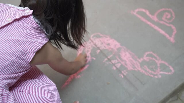 Over the shoulder view of a girl in dress drawing with a pink chalk on concrete ground