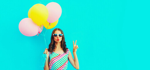 Portrait of beautiful young woman with bunch of balloons blowing her lips sends air kiss wearing a colorful dress on blue background