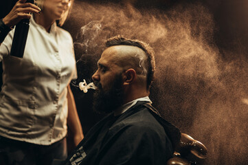 Young bearded man with a mohawk getting haircut by hairdresser and smoking a cigarette