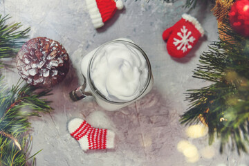 Christmas white drink eggnog on grey background with spruce branches, cone, and handmade decorative knitted ornaments sock, hat, mitten