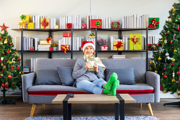 Senior Caucasian woman sitting on the sofa couch wearing red Santa hat while drinking hot chocolate with Christmas tree on the back