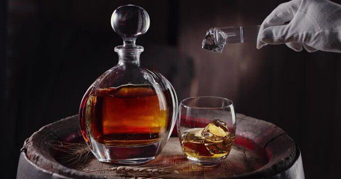 An ice cube slowly falls into a glass of whiskey on top of a vintage whiskey barrel. A decanter with a drink stands nearby, a dark brown background. Blackmagic Ursa Pro G2. 