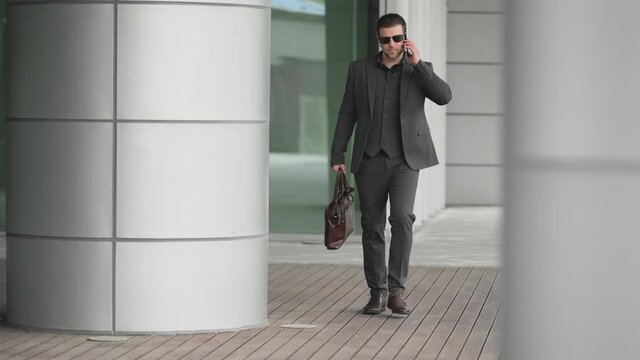 Slow motion Successful confident businessman in suit carrying a briefcase walking between office buildings  talking on the phone and getting good news make a good deal Very excited and happy