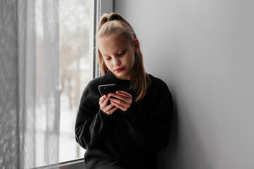 Little blonde girl in black sweater sitting on window sill and using smartphone. Bored child writing on phone indoors. Sad kid home alone during lockdown. Texting, chatting, reading.