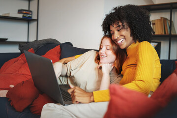 Couple of young homosexual women smiling watching content on a laptop sitting on a couch - multiracial lesbian couple in love concept - Powered by Adobe