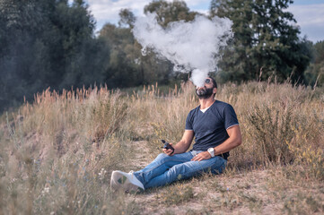 Naklejka na ściany i meble Handsome vape man enjoying an electronic smoke device on the grass. Non-tobacco smoking.