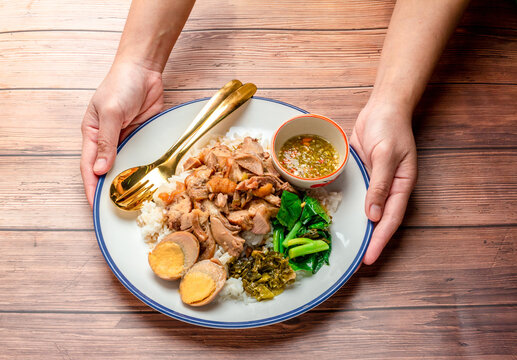 Hand holding plate of Stewed pork leg with boiled egg and vegetable sush as Kale and pickled mustard green and sour sauce place on table wooden background, Popular food in Asia.