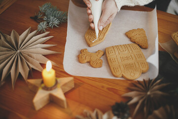 Hands decorating christmas gingerbread cookies with icing on wooden table with candle and...