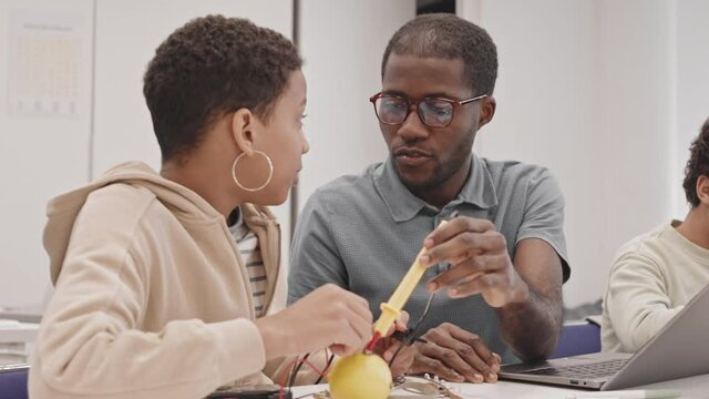 Waist-up of young Black teacher and female secondary school student sitting at table in classroom, doing experiment with electricity and lemon, talking