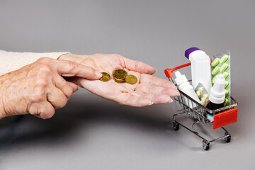 Poverty. Hands of elderly woman counting a coinson near full of medicines little shopping cart. Gray background. Concept of online shopping