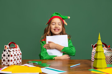 Little smiling girl dressed like funny gnome or elf sitting at table and writing letter to Santa Claus isolated over green background.