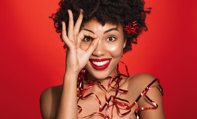 Image of young african woman with clear glowing skin, wrapped in Christmas gift ribbons and smiling, showing okay, ok sign, standing over red background