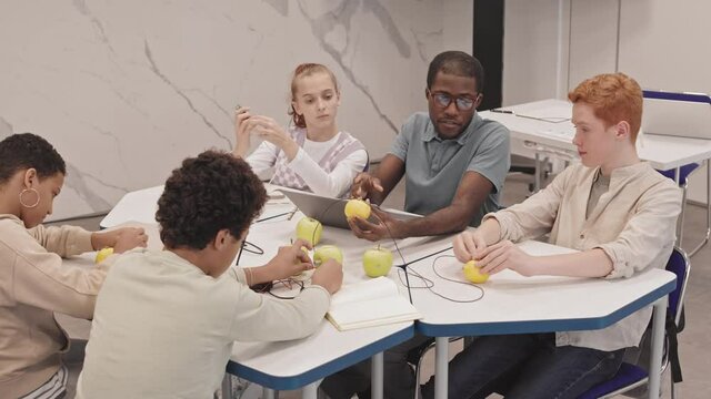 High angle of young Black male teacher and diverse secondary school students sitting at table in classroom, having electrodynamics experiment of getting electricity from fruit