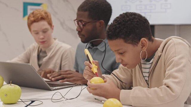 Chest-up of short-haired Black girl experimenting with lemon in physics class sitting at table with young male teacher and red-haired Caucasian boy