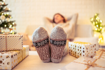 Woman sitting on couch near christmas tree. Female legs, feet in winter woolen socks putting on the...