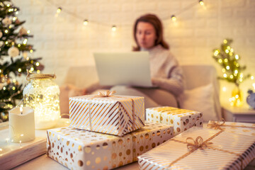 Unfocused, blur woman on couch using laptop for searching gift ideas sitting near christmas tree and packing handmade gift boxes  putting it on the table. Presents for family.