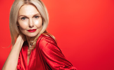Fototapeta premium Stunning elegant woman, 55 years old, touches her healthy glowing hair and looking sensual at camera, wearing red dress, makeup for new year party, st valentines day, red background