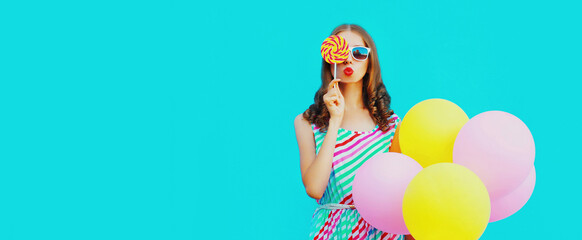 Portrait of beautiful young woman with bunch of balloons blowing her lips sends air kiss wearing a colorful dress on blue background