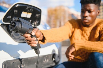 Close up of african american man connecting charging cable to electric car. Young male standing near his modern auto with leather suitcase in hand.