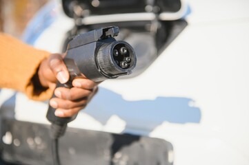 Close up of male hand holding electric charging plug for luxury red car. African businessman charging his auto on station. Eco friendly transport.