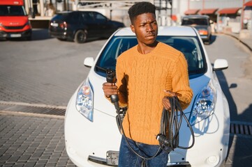 african man holding charge cable in on hand standing near luxury electric car.