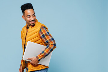 Side profile view young black man 20s years old wears yellow waistcoat shirt hold use work on laptop pc computer walking looking behind isolated on plain pastel light blue background studio portrait.