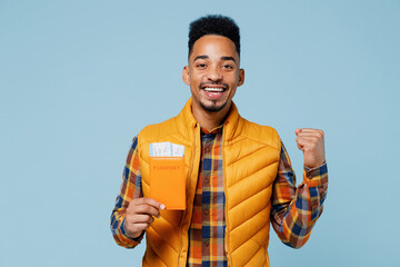Young black man 20s years old wears yellow waistcoat shirt hold passport boarding tickets doing winner gesture clenching fists say yes isolated on plain pastel light blue background studio portrait.