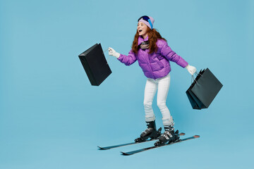 Full body happy skier woman in warm purple padded windbreaker jacket ski goggles mask spend weekend in mountains holding package bags with purchases shopping isolated on plain blue background studio.