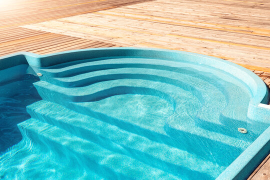 New modern fiberglass plastic swimming pool entrance step with clean fresh refreshing blue water on bright hot summer day at yard or resort hotel spa area. Wooden flooring deck of teak or larch board
