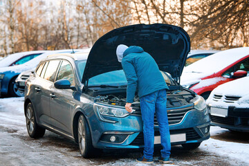 Man stands under open hood of modern car, checking engine oil level in winter season on parking...