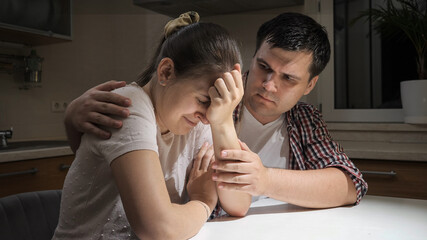 Young man consoling and hugging his crying wife on kitchen at night. Concept of despair, stress, depression, anxiety, family problems, divorce and frustration.