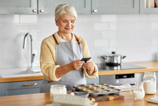 cooking, food and culinary concept - happy smiling senior woman with smartphone taking picture of cupcakes in baking mold on kitchen at home