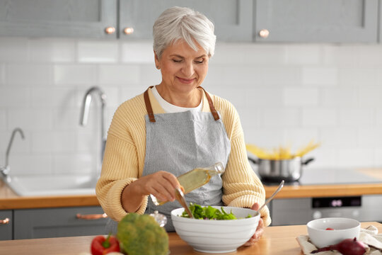 healthy eating, food cooking and culinary concept - happy smiling senior woman with olive oil making vegetable salad on kitchen at home