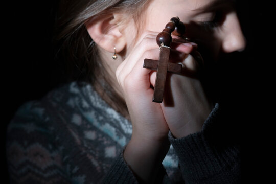Portrait of young beautiful girl praying to god with wooden rosary. Selective focus on cross. Horizontal image.