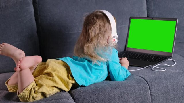 child 2-3 years old, girl in headphones lies on sofa in front of laptop, uses Internet, watches educational cartoons, plays computer games, green blank screen for video, concept of remote learning