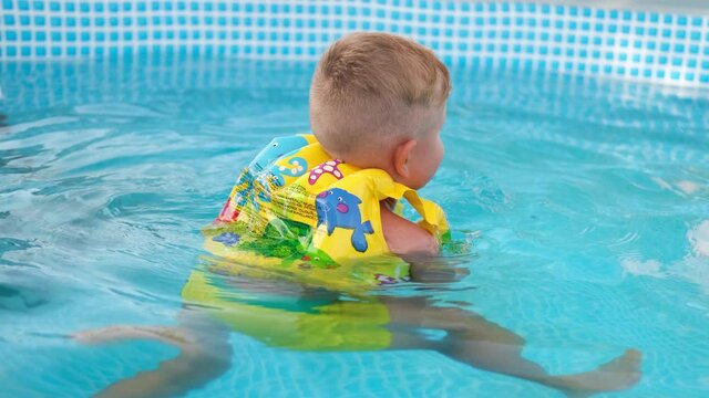 The child swims in the pool in a yellow vest. A little boy swims in a blue pool and plays with a toy. Summertime 
