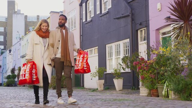 Multi-cultural couple arm in arm as they walk along cobbled mews street on visit to city in autumn or winter carrying sale shopping bags - shot in slow motion