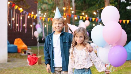 Fototapeta premium Close up of the caucasian little cute girl in a conus standing with balloons and smiling while embracing with her friend at her birthday party. Portrait